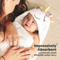 A baby wrapped in a hooded unicorn towel looks up at an adult, both lying on a beige textured surface. Text on the image reads, Impressively Absorbent. Luxuriously plush 500GSM cotton terry.