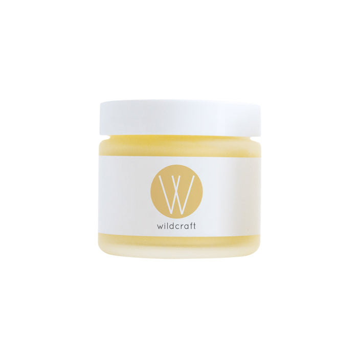 A small glass jar with a white lid and a white label featuring a gold circle with a W and the word wildcraft underneath, containing a yellow cream or balm.