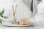Two wooden toothbrushes in a glass jar sit on a white dish with soap in a Peach Soap Saver Bag. The setting features a marble countertop, white tiled backsplash, and gray cloth. A green plant on the left adds freshness to the scene.