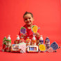 A young child in a red shirt sits smiling behind a colorful display of bath products and playful fruit character cutouts, all set against a bright red background.