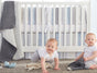 Two babies in white onesies are in front of a white crib with the Coyuchi Cozy Cotton Organic Baby Blanket, a soft gray and white accent, hanging on the side. One baby is crawling and smiling during tummy time while the other sits holding a stuffed toy. Plush toys are scattered across the carpeted floor.