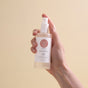 A hand holds a transparent spray bottle of Wildcraft Balance Toner, featuring a white label with a pink logo, set against a plain beige background.