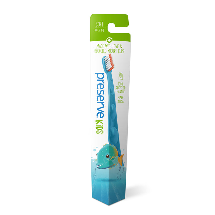 A blue kids’ toothbrush with an orange and white brush head is packaged in a green and white box labeled “Preserve Kids.” The packaging features a cartoon fish design and says its made from recycled yogurt cups.