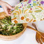 A hand covers a wooden bowl of salad with a Bees Wrap Assorted Wraps- 5 pack, featuring colorful produce prints. A wooden salad server nearby shows the practical charm of these washable, reusable food wraps for daily use.