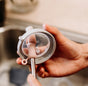 Using the pink silicone brush from Haakaas Double-Ended Silicone Cleaning & Bottle Brush set, a person cleans a clear lid with a gray rim and spout over the sink, subtly highlighting kitchen hygiene essentials for spotless baby gear and other items.