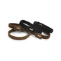 Four fabric wristbands, two brown and two black, each featuring a small rectangular patch with two metal rivets, are arranged in a casual overlapping layout on a white background.