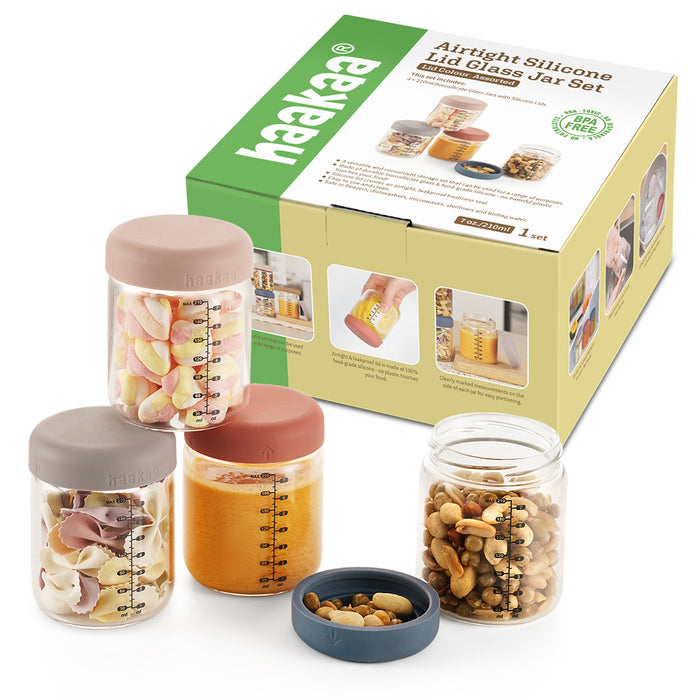 Three glass jars with colored silicone lids hold snacks and puree, placed in front of a green and beige Haakaa Airtight Silicone Lid Glass Jar Set box, with one lid open showing nuts inside the jar.