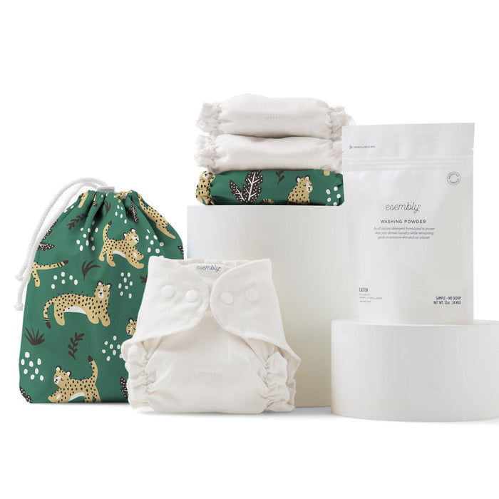 A set of reusable cloth diapers, a green drawstring bag with cheetah prints, stacked white inserts, and a pouch of washing powder are displayed against a white background.