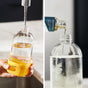 Split image: On the left, water is being poured into a glass bottle with a yellow base labeled GLASS. On the right, liquid from a small bottle is being poured into a similar clear glass bottle.