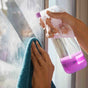 A person cleans a window with a turquoise cloth and a spray bottle labeled glass with a pink base, spraying cleaning solution on the glass.