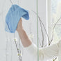 A person wearing a white sleeve uses Grove Co.s blue Daily Shower Cleaner Concentrate cloth with a plant-based formula to clean a glass surface. White flowers and thin branches are visible outside, as natural light streams through the window.