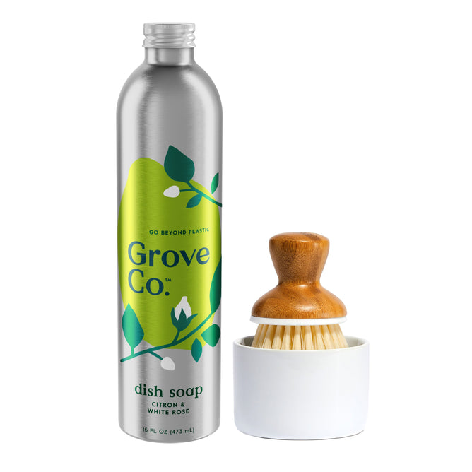The Grove Co. Dish Washing Starter Set features a silver bottle of Ultimate Dish Soap with a green Citron & White Rose label. Alongside, a wooden dish brush sits in a white holder, decorated with green leaf graphics to emphasize: go beyond plastic waste.