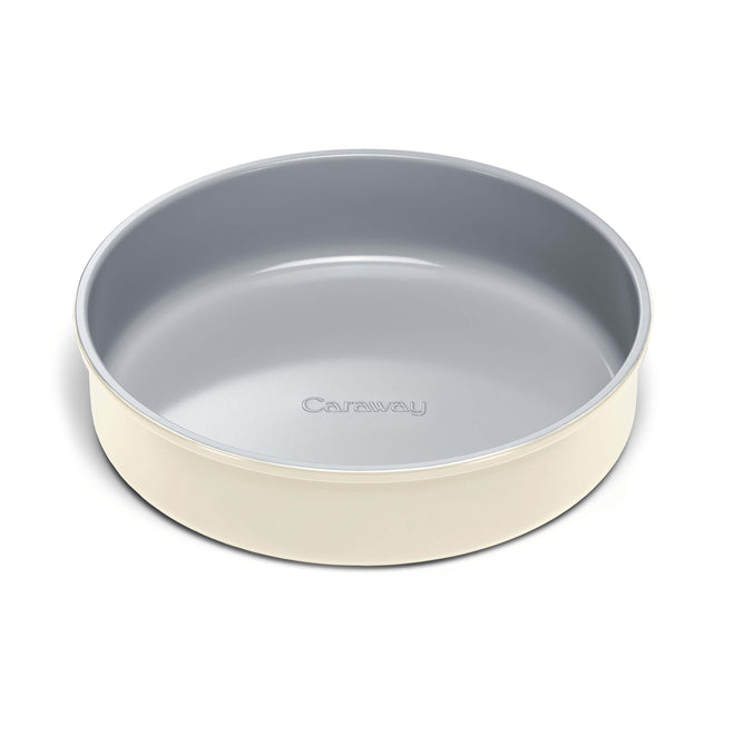 A round, light gray nonstick baking pan with high sides and a cream-colored exterior, featuring the brand name Caraway embossed on the inner bottom.