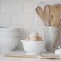 On a white kitchen counter, a bowl of eggs, stacked bowls, and various utensils including a whisk and wooden spoons are in a speckled ceramic holder. A Give it a Rest Spatula by Bambu rests near some garlic, enhancing the display of organic kitchen tools.