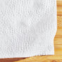 A close-up of Grove Co.s Tree-Free Napkins - 100% Bamboo shows a textured, floral, and dotted white paper towel made from FSC-certified bamboo fibers. The partially folded napkin on the wooden surface highlights its eco-friendly elegance.
