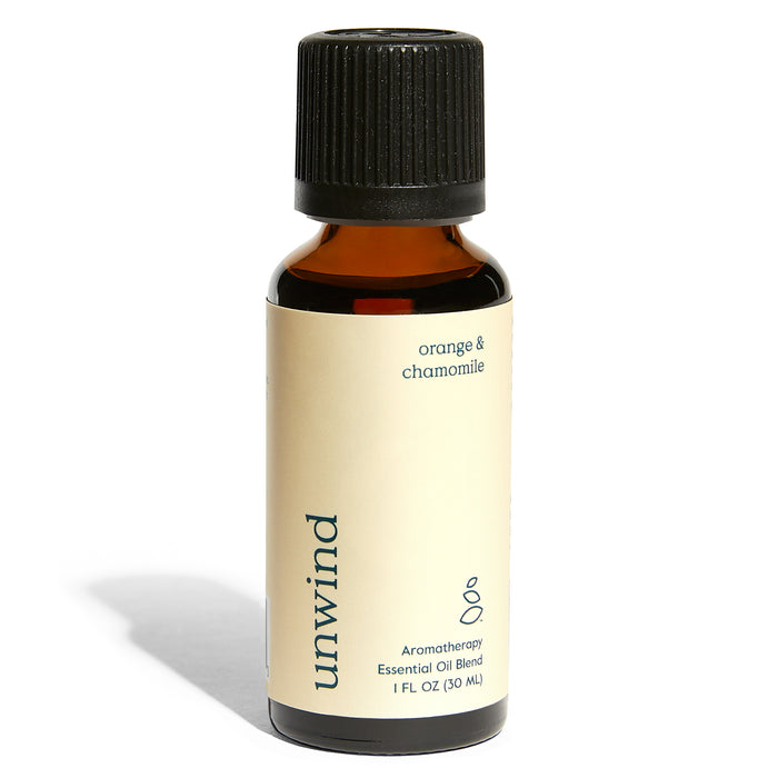 A small amber bottle labeled unwind contains orange and chamomile aromatherapy essential oil blend. The label is cream-colored with black text and a black screw-on cap. The bottle casts a shadow on a white surface.