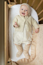 A smiling baby wearing a cream-colored organic cashmere cardigan, bonnet, and knit socks lies in a wooden crib next to a matching Cashmere Cloud Baby Blanket Gift Set - Butter Cream.