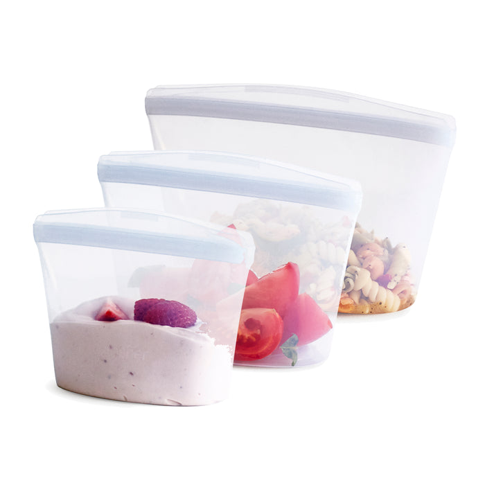 Three transparent reusable silicone food storage bags of different sizes, each containing food items: the smallest has yogurt and berries, the medium has chopped tomatoes, and the largest has mixed pasta salad.