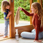 A woman kneels indoors, holding a striped cloth and smiling at a child standing beside a sliding glass door. A bottle of Mrs. Meyer’s Glass Cleaner sits on the floor nearby, ready to leave glass surfaces sparkling. Green trees are visible outside.