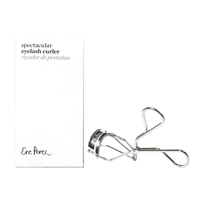 A silver eyelash curler is placed next to a white box labeled spectacular eyelash curler, rizador de pestañas with Ere Perez printed at the bottom. The background is white.