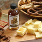 A bottle of Runamok Smoked Maple Syrups - Cheese Pairing Collection sits on a wooden board next to cubes of cheese, ideal for pairing with pretzels and snacks in the background.