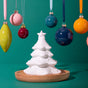 A white, sculpted Passive Diffuser, Christmas Tree sits on a wooden base, surrounded by colorful hanging ornaments.