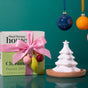 A white ceramic Passive Diffuser, Christmas Tree rests on a small wooden tray next to a gift-wrapped box adorned with a pink ribbon and small decorative ornaments.