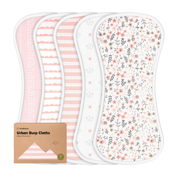 Five cotton baby burp cloths in pink, white, and floral patterns are displayed side by side. A small brown cardboard box labeled Urban Burp Cloths is positioned in front of the cloths.