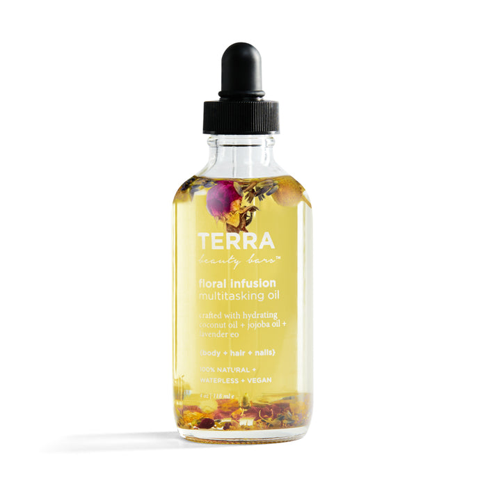 A clear glass dropper bottle labeled TERRA beauty bars floral infusion multitasking oil, containing a light yellow oil with visible dried flowers and herbs inside. The oil is for body, hair, and nails.