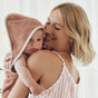A woman smiles while holding a baby wrapped in the Coyuchi Cloud Loom™ Organic Baby Hooded Towel and Bath Mitt Set. The baby looks at the camera, bathed in soft natural light.