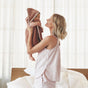 A woman in striped pajamas holds a smiling baby wrapped in the Coyuchi Cloud Loom™ Organic Baby Hooded Towel and Bath Mitt Set in a bright bedroom with white curtains and a plant in the corner.