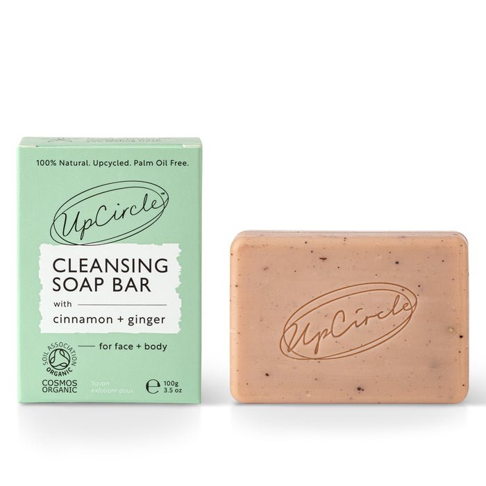 A green box labeled UpCircle Cleansing Soap Bar with cinnamon + ginger stands next to a pinkish soap bar engraved with the UpCircle logo. The products are for face and body and are organic and palm oil free.