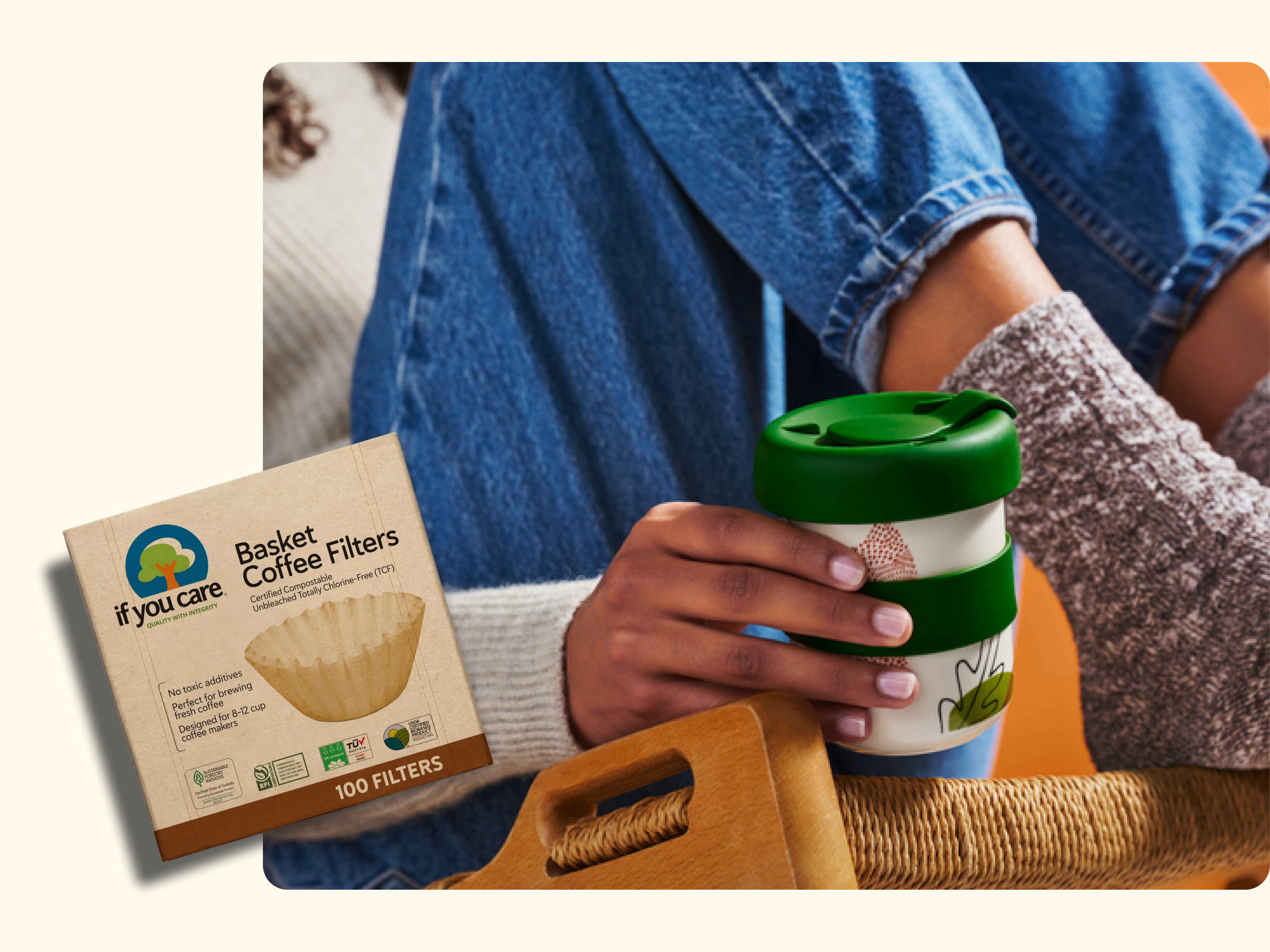 A person in jeans holds a reusable coffee cup with a green lid. Next to them is a box of If You Care basket coffee filters made from unbleached paper. A wooden basket is also visible in the foreground.