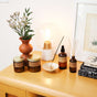 A wooden table displays a vase with flowers, a lit bulb lamp, two amber glass jars with gold lids, a spray bottle, an incense stick burning in a dish, a reed diffuser, and a stack of books.