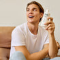 A smiling person in a white t-shirt sits on a couch, holding up a bottle of Phil’s Strengthening + Thickening Conditioner for thinning hair. The background is simple and light-colored.