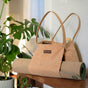 A Yoloha Yoga Cork Tote Bag, designed with a built-in yoga mat holder and a compartment for a water bottle, rests on a wooden shelf next to a green monstera plant in a bright, modern room.