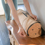 A person in light blue leggings unzips the Yoloha Yoga Cork Yoga Mat Carrier on a wooden bench, with natural light streaming in from a nearby window.