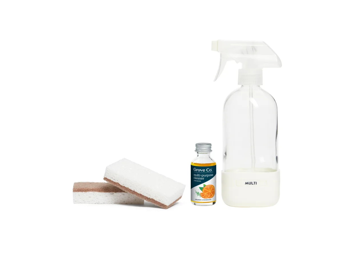 A spray bottle labeled “MULTI,” a small bottle of cleaner with an orange on the label, and two white cleaning sponges with brown scrubber sides are arranged on a white background.