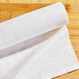 A close-up of a single white paper towel partially unrolled on a light wooden surface. The towel has a soft texture and casts a slight shadow, with the wood grain visible underneath.