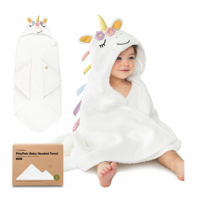 A smiling baby sits wrapped in a white hooded towel designed as a unicorn, with a yellow horn, colorful ears and mane. The towel package and a folded towel are also shown nearby.