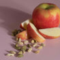 A red and yellow apple sits on a pink surface with several slices cut from it. Cardamom pods are scattered next to the apple and slices.