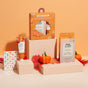 Skincare products and gift boxes with pumpkin-themed packaging are displayed on beige blocks, surrounded by autumn leaves, small pumpkins, and a pumpkin print card, set against a warm peach background.