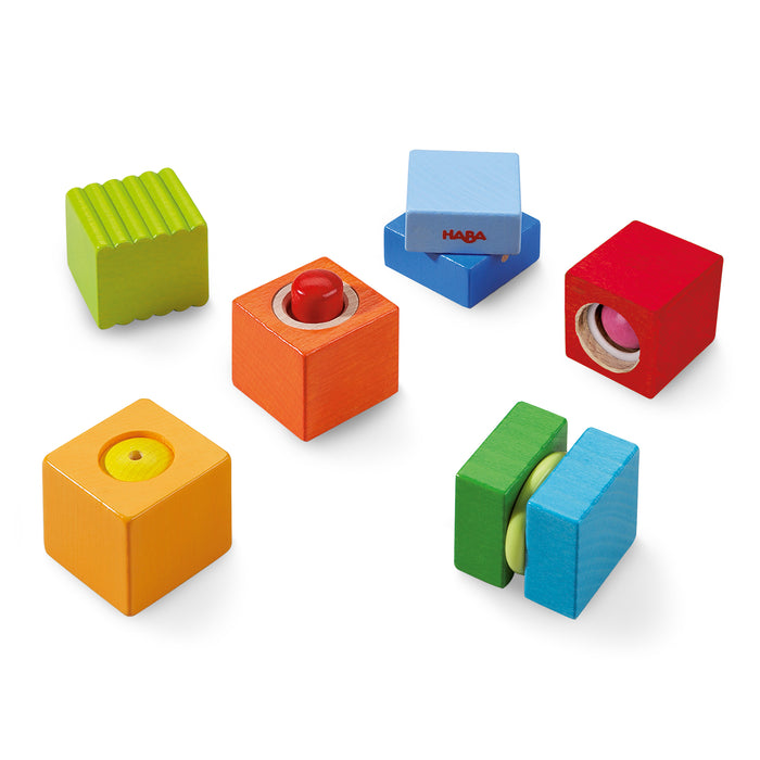 Six colorful wooden sensory blocks in different colors and textures, including red, blue, yellow, green, and orange, each with unique features like buttons, ridges, and spinning parts, arranged on a white background.