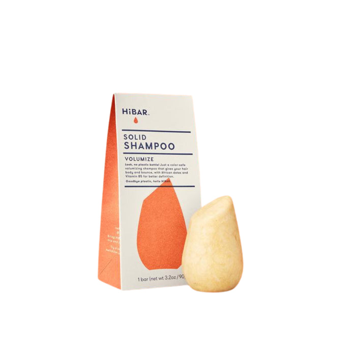 A HiBAR Solid Shampoo Volumize bar and its packaging box are shown. The box is upright with text and an orange design; the yellowish, teardrop-shaped shampoo bar stands in front of it.