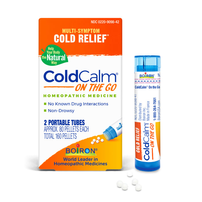 Box and portable tube of ColdCalm On The Go homeopathic cold relief medicine by Boiron. The box highlights drug-free, non-drowsy, and natural ingredients, with small white pellets scattered nearby.