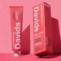 A pink tube and matching box of Davids Kids + Adults Premium Toothpaste with white lettering, shown against a pink backdrop. The packaging emphasizes that it is a fluoride-free toothpaste crafted with natural ingredients and offers a tasty strawberry flavor suitable for both children and adults.