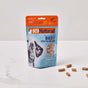 A standing pouch of K9 Natural Beef Healthy Bites dog treats is displayed on a white surface, with several treat pieces scattered beside it. The package features an image of a dogs face and product information.