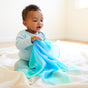 A baby in a light blue outfit sits on a soft white surface, holding Sarahs Silks Silk and FSC-Certified Wood Baby Teether. The room is bright with white walls and floor. The baby gazes ahead with curiosity, clutching the blue-green sheer fabric.