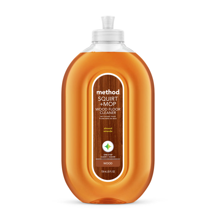 A bottle of Method Squirt + Mop Wood Floor Cleaner in almond scent, featuring a clear, curvy bottle filled with amber-colored liquid and a label describing the product details.