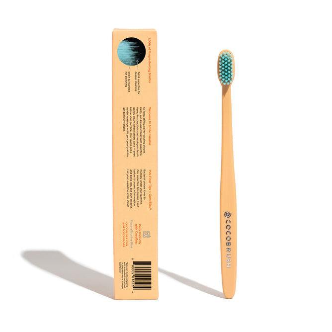 A bamboo toothbrush with turquoise bristles stands upright next to its brown rectangular packaging, which displays product information and a barcode. The package has a circular cutout showing the bristles.